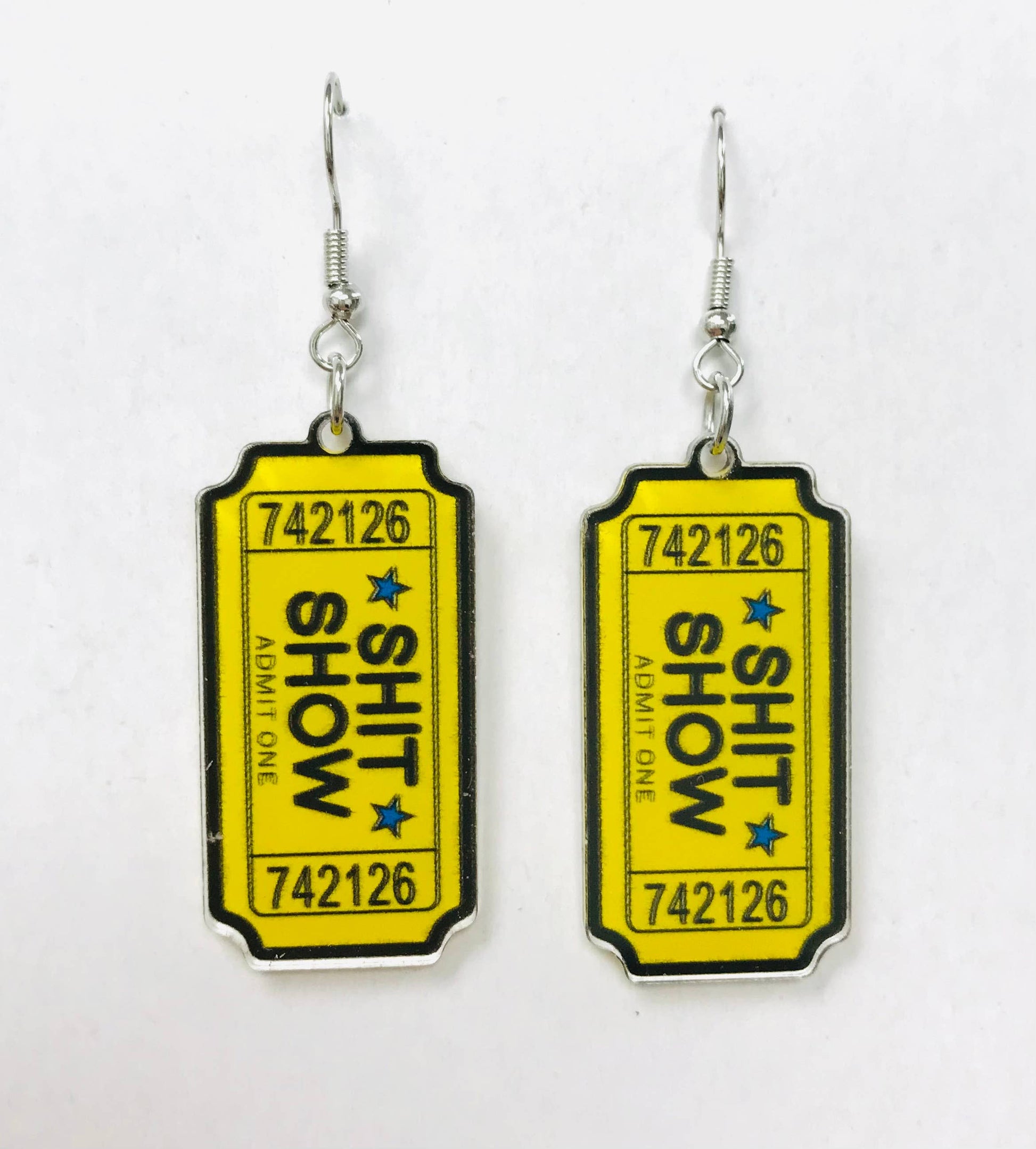 Acrylic earrings shaped like yellow admission tickets with "SHIT SHOW" text and blue stars.