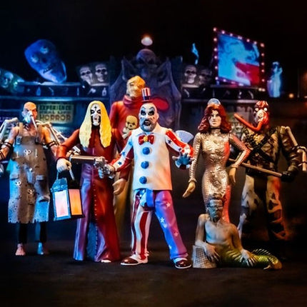 A group of horror-themed action figures or collectible characters posed together on a dark display stand. The figures include clown, zombie, monster, and mermaid-style costumes, with a dark themed backdrop featuring masks and screens.